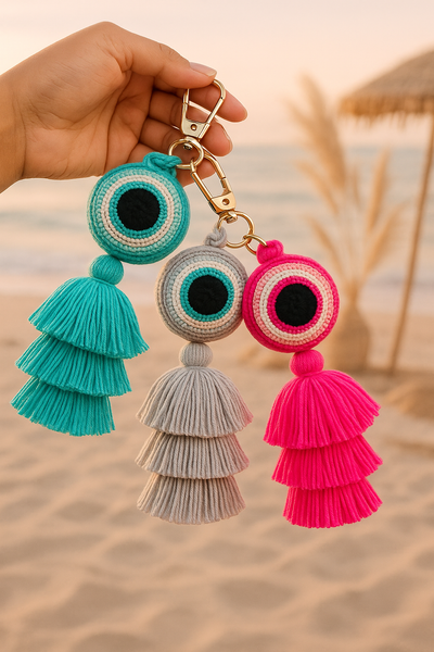 Three colorful tassel keychains held against a blurred beach background