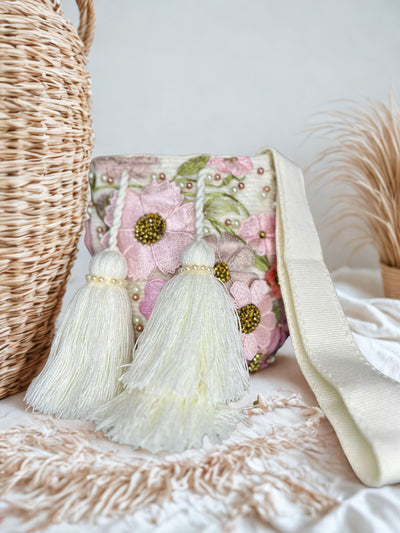 Boho chic crossbody bag with pink embroidered flowers, gold bead centers, and white pearl details, featuring two large pearl-beaded tassels. The bag has a hand-loomed strap and a white crocheted base.