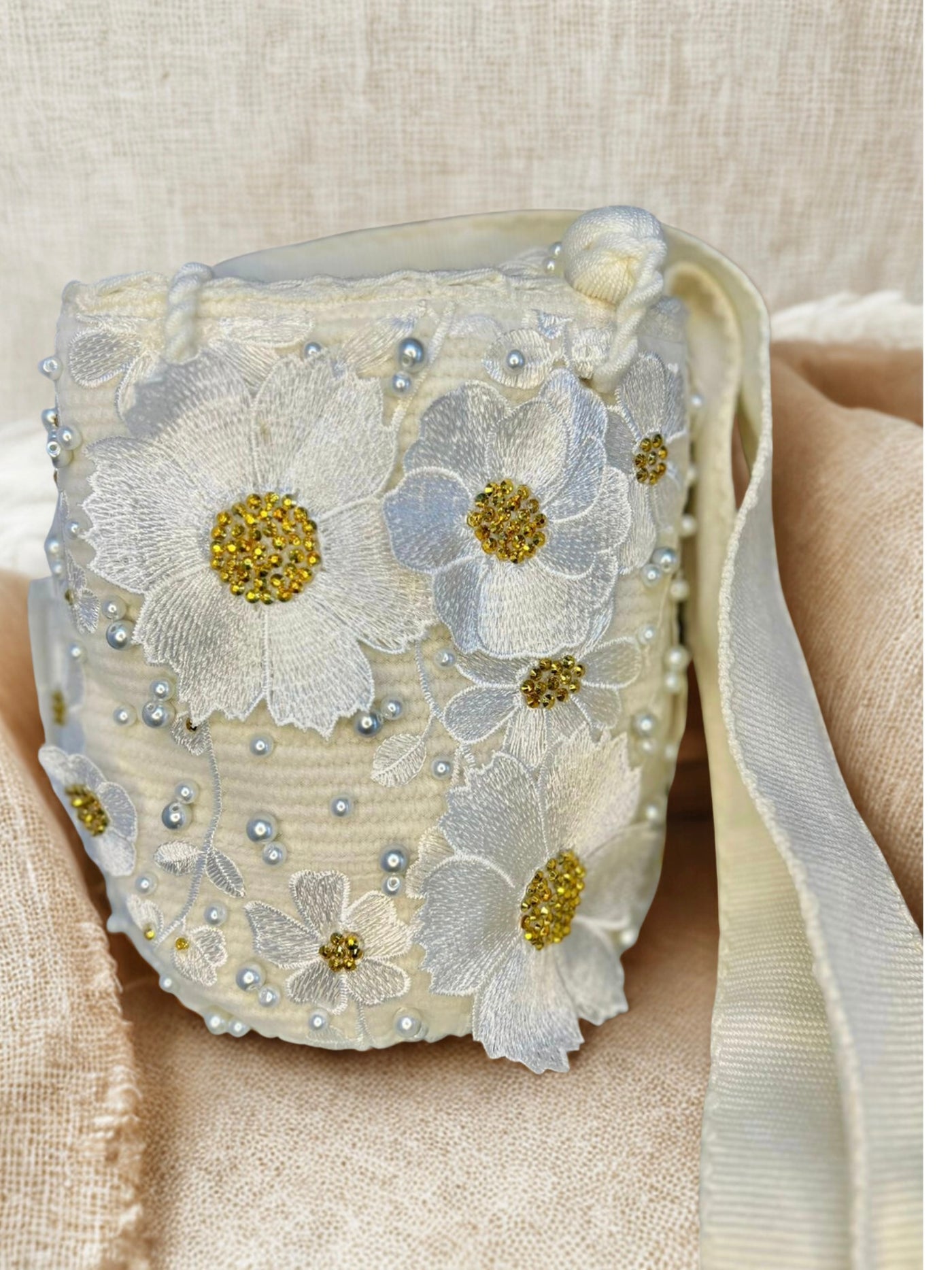 Artisan bucket bag with white embroidered flowers, gold bead centers, and scattered white pearls. The hand-crocheted bag has a wide white strap and a subtle background sheen.