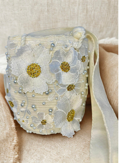 Artisan bucket bag with white embroidered flowers, gold bead centers, and scattered white pearls. The hand-crocheted bag has a wide white strap and a subtle background sheen.