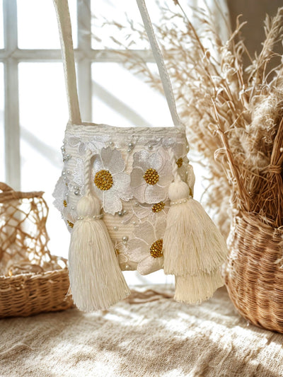 White artisan bucket bag with intricate white floral embroidery, golden beaded centers, and scattered pearls, hanging in front of a window. It has two large white tassels.