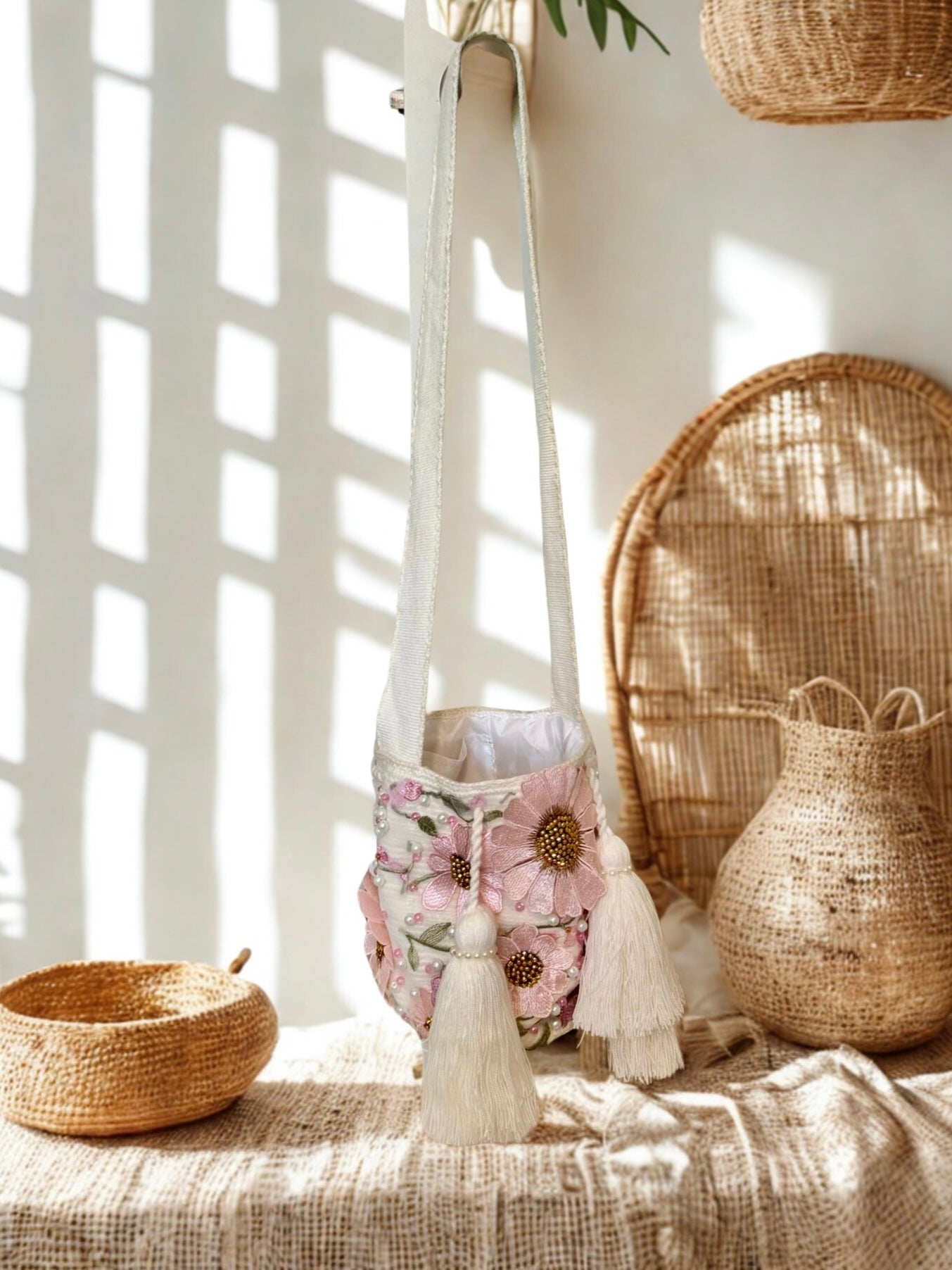 Boho chic bucket crossbody bag with pink floral embroidery, gold beads, and pearl embellishments, hanging from a white wall. It features two prominent white tassels and a hand-loomed strap.